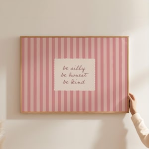 Puede incluir: Una impresión enmarcada con un fondo a rayas rosas y blancas. La impresión presenta las palabras "be silly, be honest, be kind" en un estilo manuscrito dentro de un rectángulo blanco con bordes festoneados. El marco es de color madera clara.