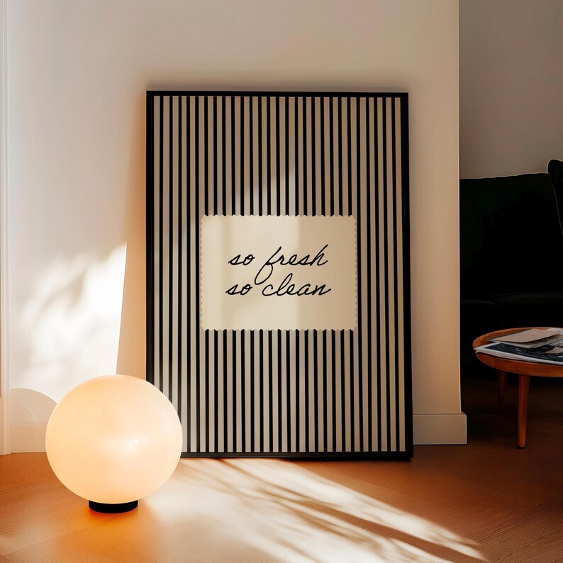 May include: A framed artwork with a black and white striped background and the text "so fresh so clean" in a cursive font. A round, glowing lamp with a black base sits in front of the artwork.