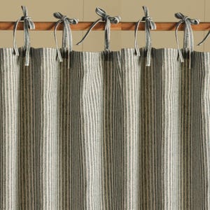 Puede incluir: Un panel de cortina a rayas en tonos grises y blancos, colgado de una barra de madera. La cortina tiene rayas verticales y está atada con lazos grises en la parte superior. La tela parece ser lino o un material natural similar.