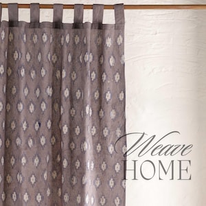 以下が含まれることがあります： 木製の棒とタッセルが付いたグレーと白のパターンのカーテン。カーテンは、壁に「Weave HOME」という文字が書かれた白い壁の前に吊るされています。