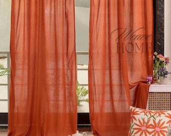 May include: Two rust-colored linen curtains with a tie-top design hang in a window. The curtains are gathered at the top and tied with a knot. The curtains are made of a lightweight, breathable fabric. The curtains are hung on a metal rod. The curtains are in a room with a wooden floor and a rug. There is a white teapot and two glasses on the rug. The curtains are a warm, inviting color and would be perfect for a living room or bedroom.