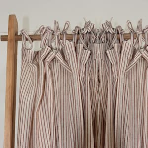 Puede incluir: Un conjunto de cortinas blancas con finas rayas rojas, atadas con lazos de tela y colgadas de una barra de madera.