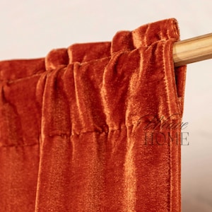 Puede incluir: Primer plano de un panel de cortina de terciopelo color óxido colgado de una varilla de madera. La tela tiene una textura suave y aterciopelada.