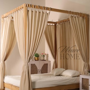 May include: A wooden four-poster bed with cream-colored curtains and a white mattress. The bed frame is made of light-colored wood. Two white pillows with pom-pom trim rest on the bed. The text "Weave HOME" is visible on the right side of the bed.