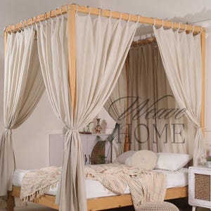 Puede incluir: Una cama con dosel de madera con cortinas color crema y una manta a juego. El marco de la cama es de madera clara y las cortinas están atadas. Un pequeño puf redondo tejido se encuentra al pie de la cama.