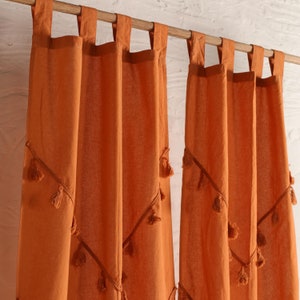 May include: Two orange cotton curtains with tassel trim hang from a wooden rod. The curtains are sheer and have a subtle diamond pattern.