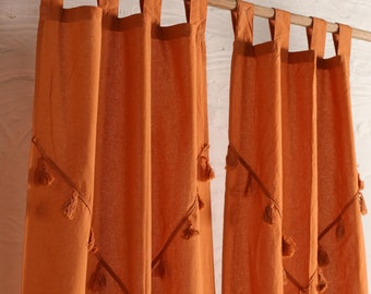 May include: Two orange cotton curtains with tassel trim hang from a wooden rod. The curtains are sheer and have a subtle diamond pattern.