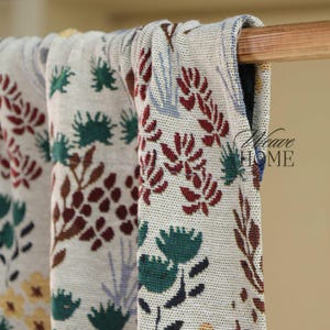 Op de afbeelding: Close-up van drie bloemmotief dekens die aan een houten roede hangen. De dekens hebben een crèmekleurige achtergrond met rode, groene, bruine en blauwe bloemmotieven. De tekst "Weave HOME" is zichtbaar.