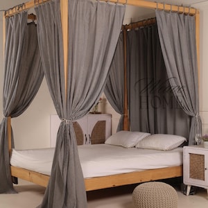 Puede incluir: Una cama con dosel de madera con un dosel de algodón a rayas grises y blancas. La cama tiene un colchón blanco y dos almohadas blancas. Un reposapiés tejido se encuentra frente a la cama.