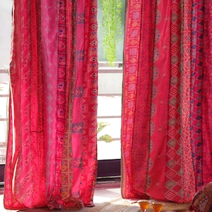 Puede incluir: Dos cortinas rojas con un patrón geométrico colorido cuelgan en una ventana. Las cortinas están hechas de una tela transparente y son ligeramente translúcidas. Las cortinas están corridas hacia atrás para revelar una vista de un patio con una alfombra y dos vasos en una bandeja.