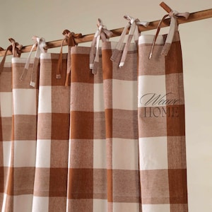 May include: Brown and white checked curtains suspended from a wooden rod. The curtains are tied with fabric bows. The text "Weave HOME" is printed on the curtain. The curtains have a farmhouse style.