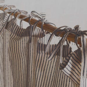 Könnte beinhalten: Gestreifte Vorhänge mit Bindebändern, die an einer Holzstange hängen. Die Vorhänge weisen vertikale Streifen in einer neutralen Farbpalette auf, wobei die Bindebänder ein dekoratives Element hinzufügen. Die Gesamtästhetik ist schlicht und rustikal.