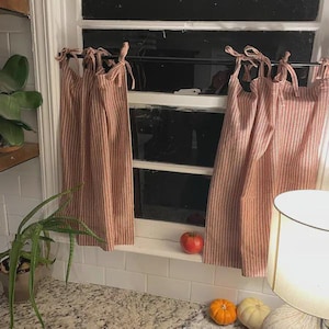 May include: Red and white striped curtains are tied back from a window. A red tomato and two small pumpkins sit on the windowsill. A potted plant is in the foreground, and a lamp is in the right corner.