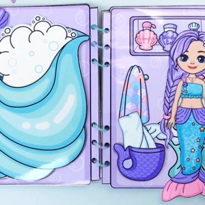 May include: A mermaid-themed paper doll playset. The open book shows a mermaid doll with a long braid, a blue top, and a shimmering tail. The background features a bathtub with bubbles and bathroom accessories.