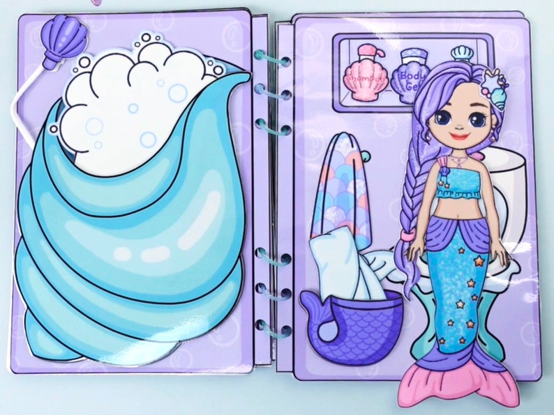 May include: A mermaid-themed paper doll playset. The open book shows a mermaid doll with a long braid, a blue top, and a shimmering tail. The background features a bathtub with bubbles and bathroom accessories.