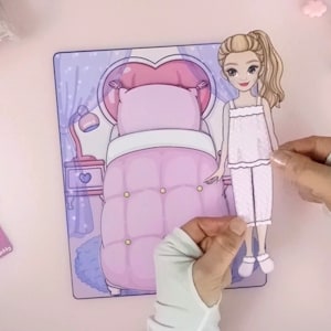 Op de afbeelding: Een papieren pop in pyjama's wordt vastgehouden in de buurt van een bijpassende slaapkamersc&egrave;ne. De slaapkamersc&egrave;ne heeft een bed met een hartvormig hoofdeinde, een nachtkastje en een lamp. Het kleurenschema is voornamelijk roze en paars.