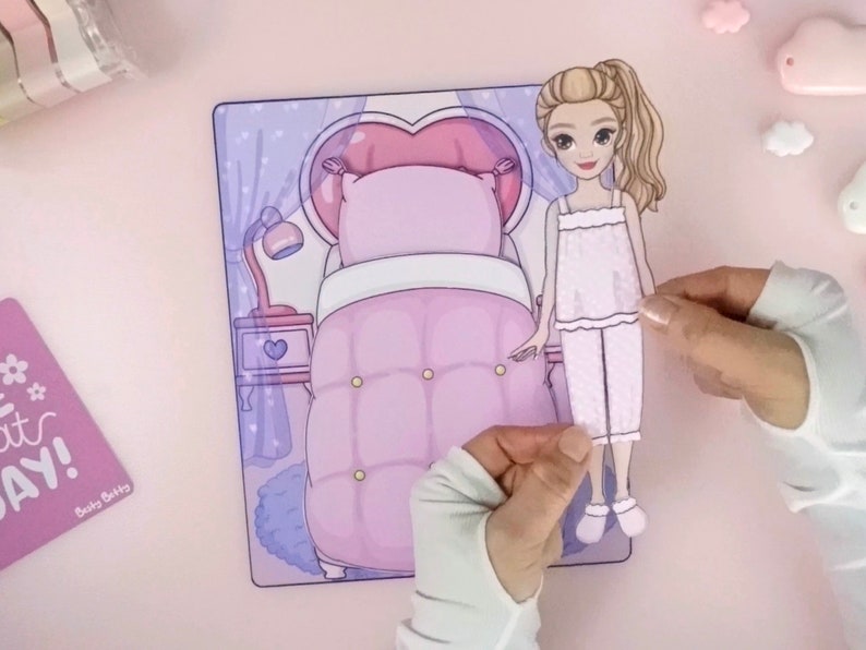 Op de afbeelding: Een papieren pop in pyjama's wordt vastgehouden in de buurt van een bijpassende slaapkamersc&egrave;ne. De slaapkamersc&egrave;ne heeft een bed met een hartvormig hoofdeinde, een nachtkastje en een lamp. Het kleurenschema is voornamelijk roze en paars.
