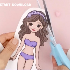 May include: A paper doll cutout of a girl with brown hair, wearing a purple bikini top and bottom. The doll is being cut out with blue scissors.
