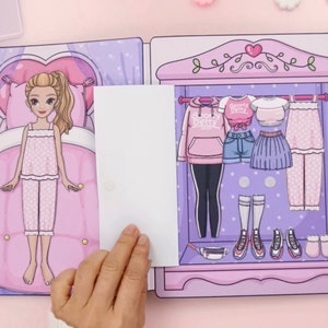 Op de afbeelding: Een papieren poppenset met een slaapkamer en een kledingkast met verschillende outfits. De pop draagt een roze pyjama. De kledingkast bevat een roze hoodie, shorts, een rok, een broek en schoenen. De tekst "Sporty Betty" is zichtbaar op een shirt.