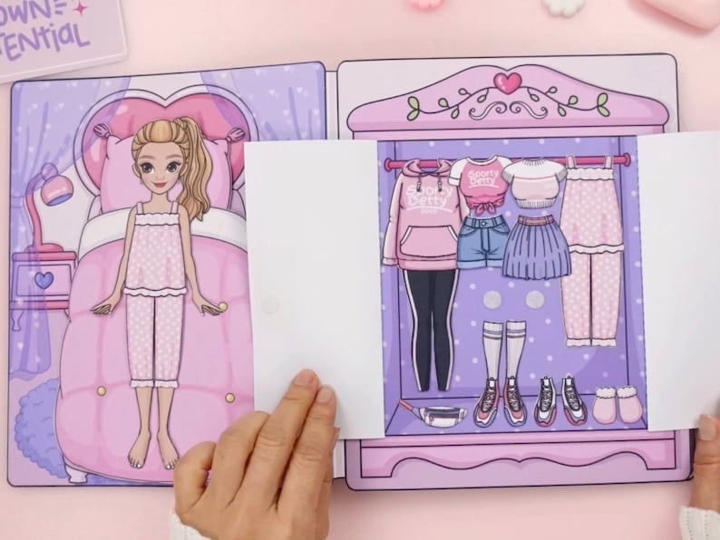 Op de afbeelding: Een papieren poppenset met een slaapkamer en een kledingkast met verschillende outfits. De pop draagt een roze pyjama. De kledingkast bevat een roze hoodie, shorts, een rok, een broek en schoenen. De tekst "Sporty Betty" is zichtbaar op een shirt.