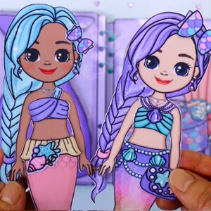 May include: Two paper doll mermaids with long hair, one blue and one purple. They wear seashell-themed tops and skirts. The dolls have bows in their hair and are holding hands. The background is a light purple.