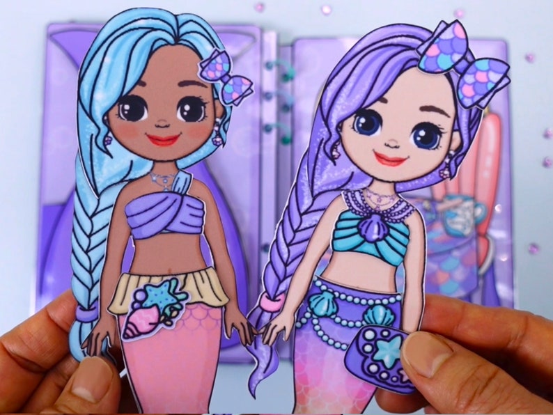 May include: Two paper doll mermaids with long hair, one blue and one purple. They wear seashell-themed tops and skirts. The dolls have bows in their hair and are holding hands. The background is a light purple.