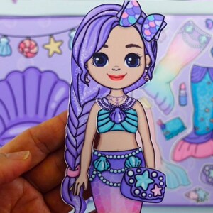 May include: A mermaid paper doll with long purple hair, a teal seashell top, and a purple mermaid tail. The doll is holding a purple purse with a star design. Accessories include a seashell, lipstick, and a mermaid tail.