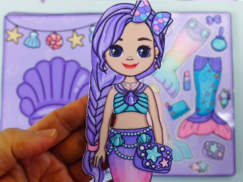 May include: A mermaid paper doll with long purple hair, a teal seashell top, and a purple mermaid tail. The doll is holding a purple purse with a star design. Accessories include a seashell, lipstick, and a mermaid tail.