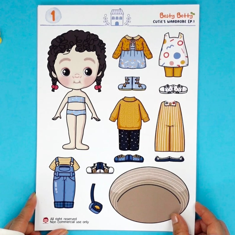 Op de afbeelding: Een papieren poppenset getiteld "Cutie's Wardrobe EP.1" met een cartoonmeisje en verschillende outfits. De outfits omvatten een gele trui, een blauwe overall en een gestreepte jas. De tekst "Besty Betty" is zichtbaar.