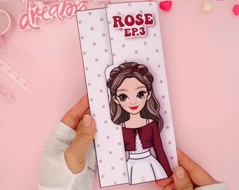 May include: A white card with a gray polka dot pattern. The card is folded in half and has a cut-out window that reveals a cartoon image of a girl with brown hair and a red shirt. The text "ROSE EP.3" is printed in red letters on the front of the card.