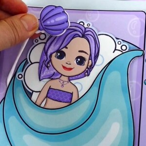 May include: A mermaid-themed paper doll with purple hair and a seashell accessory. The doll is in a blue and purple setting, with a matching seashell. The image shows a hand placing the seashell on the doll.