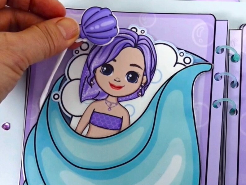 May include: A mermaid-themed paper doll with purple hair and a seashell accessory. The doll is in a blue and purple setting, with a matching seashell. The image shows a hand placing the seashell on the doll.