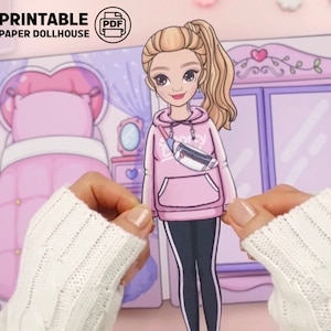 Op de afbeelding: Een afdrukbare papieren poppenhuisset. De afbeelding toont een blonde papieren pop in een roze hoodie en zwarte legging. De pop houdt een papieren uitsnede van zichzelf vast. De achtergrond bevat een roze slaapkamer met een bed en een kledingkast.