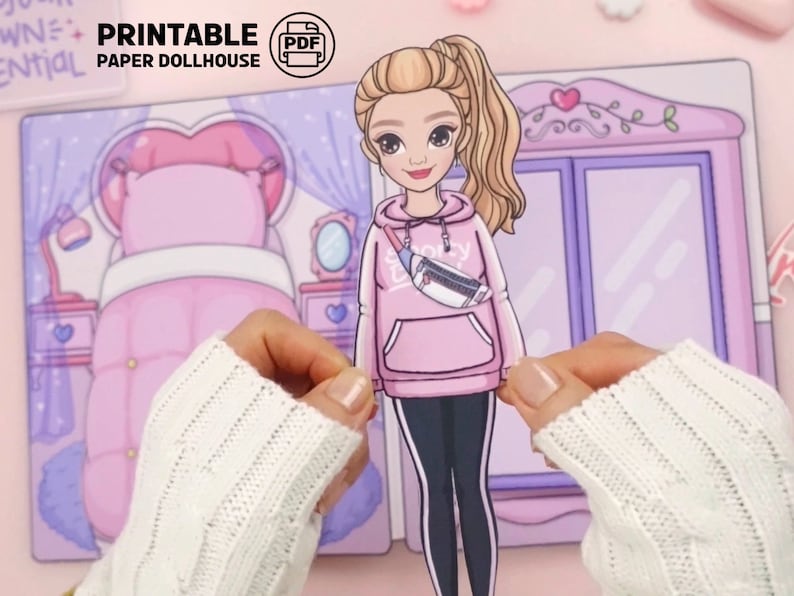 Op de afbeelding: Een afdrukbare papieren poppenhuisset. De afbeelding toont een blonde papieren pop in een roze hoodie en zwarte legging. De pop houdt een papieren uitsnede van zichzelf vast. De achtergrond bevat een roze slaapkamer met een bed en een kledingkast.