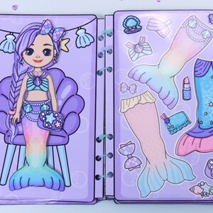 May include: A mermaid paper doll book with a purple cover. The left page features a mermaid with purple hair and a seashell top. The right page has mermaid tail options, accessories, and clothing cutouts in pastel colors. The book is bound with a spiral binding.
