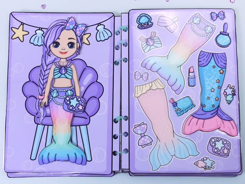 May include: A mermaid paper doll book with a purple cover. The left page features a mermaid with purple hair and a seashell top. The right page has mermaid tail options, accessories, and clothing cutouts in pastel colors. The book is bound with a spiral binding.