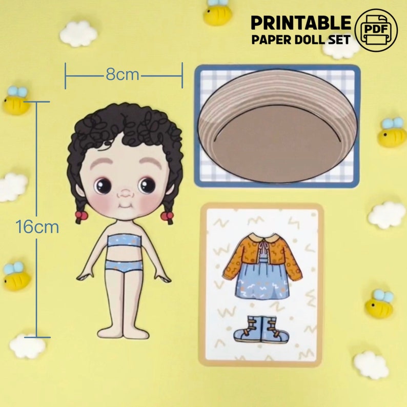 Op de afbeelding: Een printbare papieren poppenset met een pop (16 cm hoog, 8 cm breed) met krullend zwart haar en twee outfits. De set bevat een hoed en een jurk met een jasje en laarzen. De tekst "PRINTABLE PAPER DOLL SET" is zichtbaar.