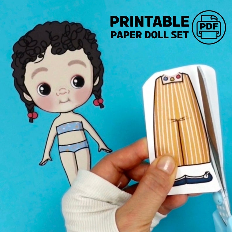 Op de afbeelding: Een afdrukbare papieren poppenset met een cartoonpop en kleding. De pop heeft donker krullend haar en draagt een blauw en wit badpak. De tekst "PRINTABLE PAPER DOLL SET" staat bovenaan. Een hand knipt een broek uit.