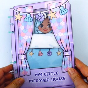 May include: A paper doll house with a mermaid theme. The cover is lavender with a mermaid illustration and the text "MY LITTLE MERMAID HOUSE". The interior features a mermaid character behind curtains decorated with seashells and stars.