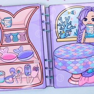 May include: A paper doll playset featuring a mermaid theme. The set includes a mermaid character with purple hair, a table, and various accessories like mermaid tails, mugs, and lollipops. The color palette is primarily pastel purples, pinks, and blues.