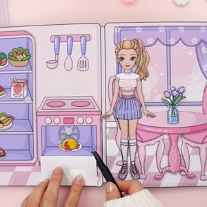 Op de afbeelding: Een papieren poppenset met een roze keukensc&egrave;ne en een poppenfiguur. De keuken heeft een koelkast, een fornuis en een tafel. De pop draagt een crop top en rok. De set bevat eten en keukengerei.