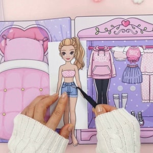Op de afbeelding: Een papieren pop met een garderobe vol outfits. De pop heeft blond haar en draagt een roze en witte tube top en een spijkerbroek. De garderobe bevat een roze hoodie, rok en pyjama. De achtergrond is een slaapkamer met een bed en een lamp.