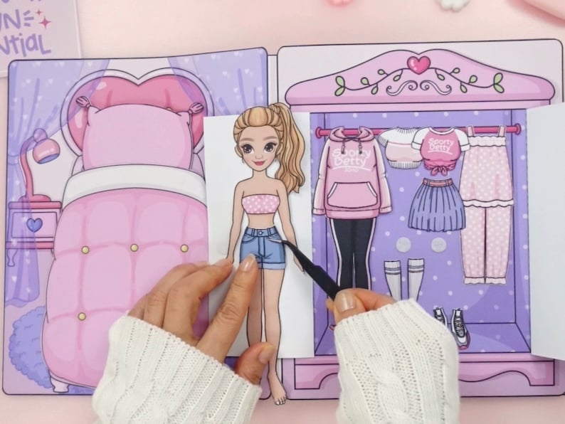 Op de afbeelding: Een papieren pop met een garderobe vol outfits. De pop heeft blond haar en draagt een roze en witte tube top en een spijkerbroek. De garderobe bevat een roze hoodie, rok en pyjama. De achtergrond is een slaapkamer met een bed en een lamp.