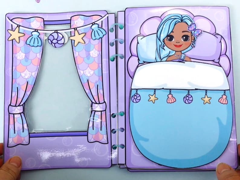 May include: A paper doll playset featuring a mermaid theme. The open book shows a bedroom scene with a mermaid doll in bed, and a window with curtains. The curtains and bedspread are light blue, and the room is decorated with seashell and star ornaments.