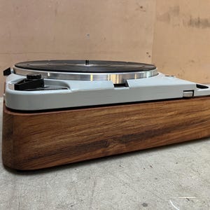 May include: A vintage turntable with a light gray body and a dark wood base. The turntable features a black platter and control knobs. The wooden base has a rich, dark brown color and a smooth finish. The design is a blend of retro and modern aesthetics.