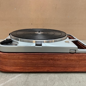 May include: A vintage turntable with a black record on a light gray base. The base is mounted on a rectangular, reddish-brown wooden plinth. The turntable features a silver tone arm and controls.