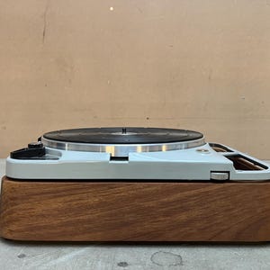 May include: A vintage turntable with a black record on a light gray base, set on a dark wood plinth. The turntable features a silver tone arm and controls. The design is minimalist and retro.