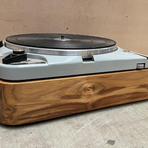 May include: A vintage turntable with a light gray body and a black record. The base is made of natural wood with visible grain. The turntable is designed for playing vinyl records.