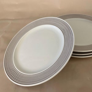 May include: A stack of white oval serving platters with a dark gray striped border. The plates are stacked on a neutral background. The plates are a simple, elegant design.