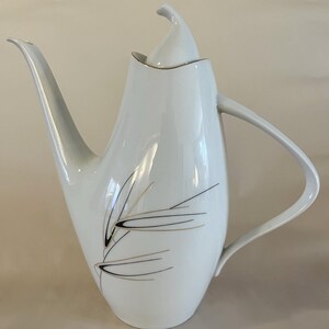 Op de afbeelding: Witte theepot van keramiek met een gouden rand en een gestileerd bladontwerp in zwart en goud. De theepot heeft een deksel met een puntige bovenkant.